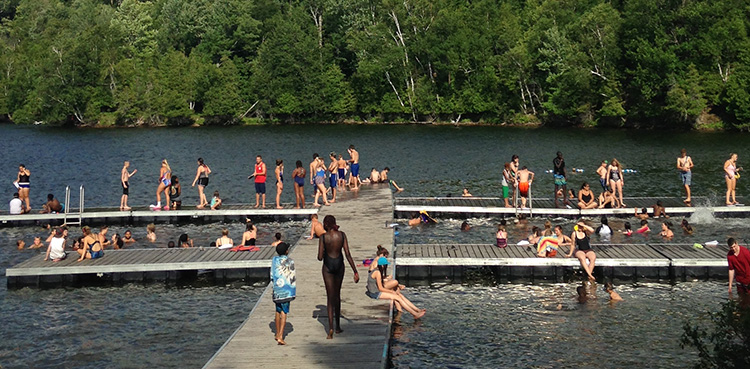 large dock on the lake with many camp members sitting on it or swimming in the water